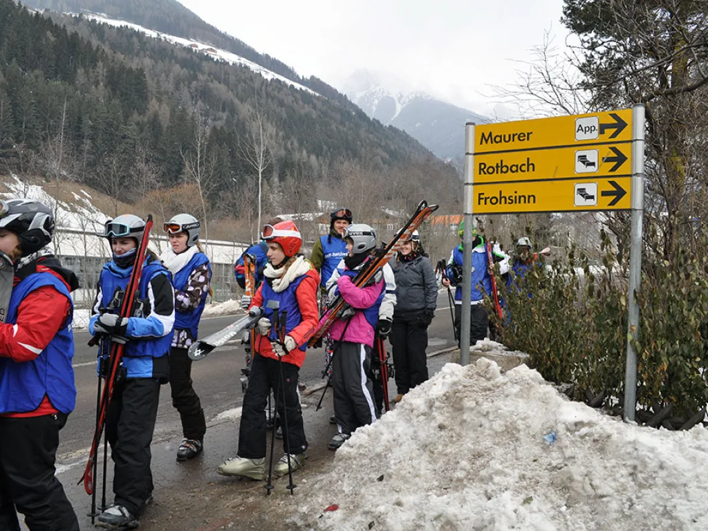 Gruppe geht mit Skiern an einem Wegweiser vorbei zur Piste.